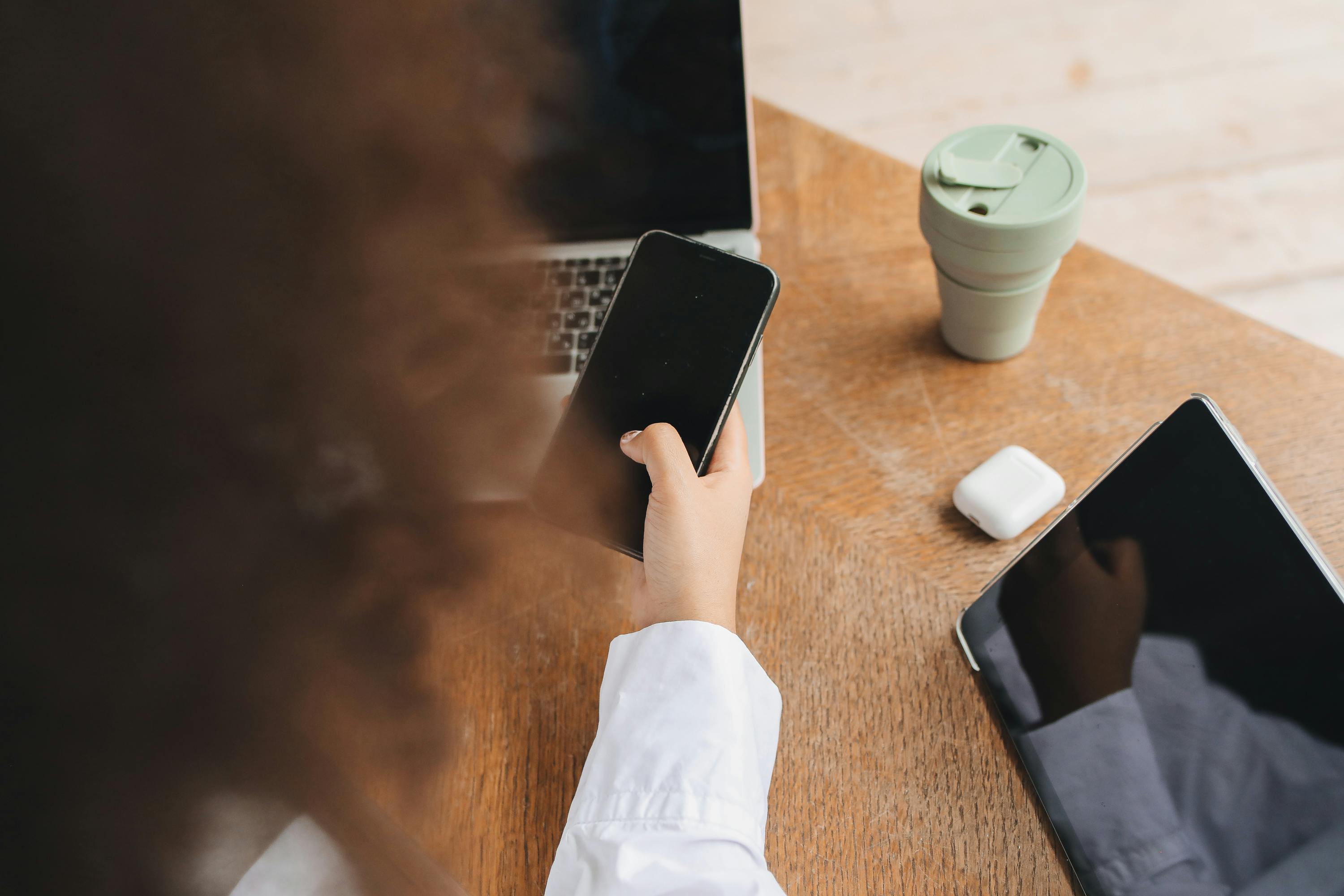 arm holding mobile phone, beside a lap top and tablet, on desk